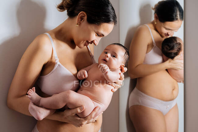 Entzückte Frau in Unterwäsche kuschelt nacktes Baby neben Spiegel, während sie sich an Wand lehnt und lächelt — Stockfoto
