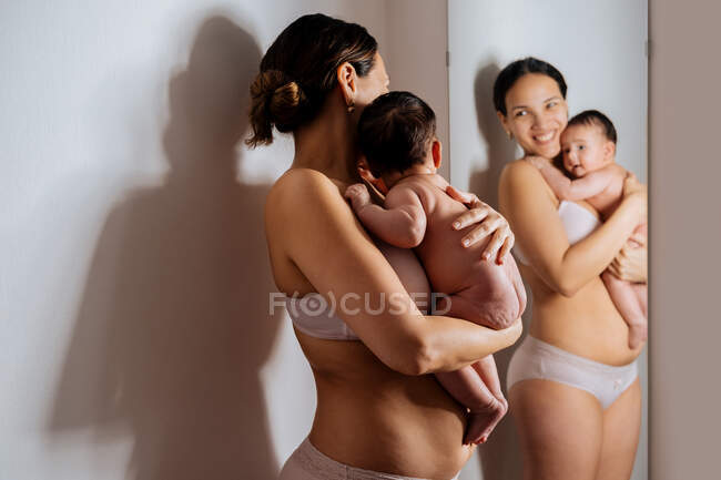 Восхитительная женщина в нижнем белье обнимает обнаженного ребенка возле зеркала, опираясь на стену и улыбаясь — стоковое фото