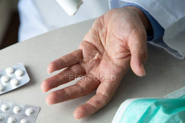 Draufsicht des männlichen Therapeuten in medizinischem Gewand, der am Tisch im Krankenhaus sitzt und Hände mit Antiseptikum desinfiziert, während er sich auf die Behandlung von Patienten während des Coronavirus-Ausbruchs vorbereitet — Stockfoto