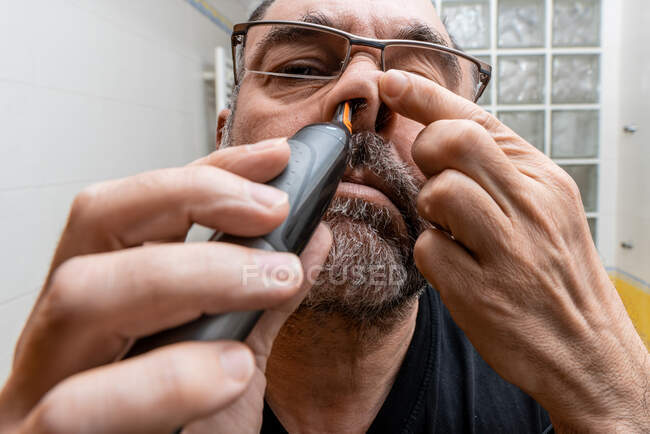 Low angle of middle aged bearded male standing in front of mirror and removing hair from nose with trimmer during morning procedure in bathroom — Stock Photo