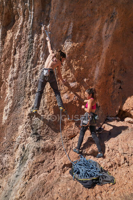 Desde abajo vista de la mujer ayudando sin camisa amigo alpinista masculino mientras ascendente y de pie con la cuerda en la mano en el suelo rocoso cerca de la montaña - foto de stock