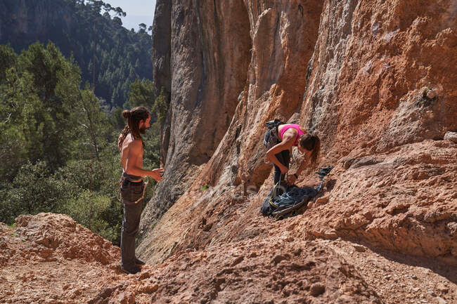 Вид збоку невідомих мандрівників, що стоять на кам'янистому ґрунті біля кам'яного схилу гори і готують мотузку та інше обладнання для скелелазіння — стокове фото