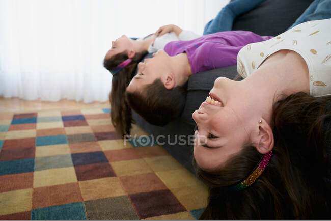 Side view of group of smiling siblings in casual clothes hanging their heads of sofa to floor while spending time together at home — Stock Photo