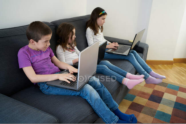 Grupo de crianças sérias em jeans casuais e camisas se reunindo em sofá confortável na sala de estar e laptops de navegação — Fotografia de Stock