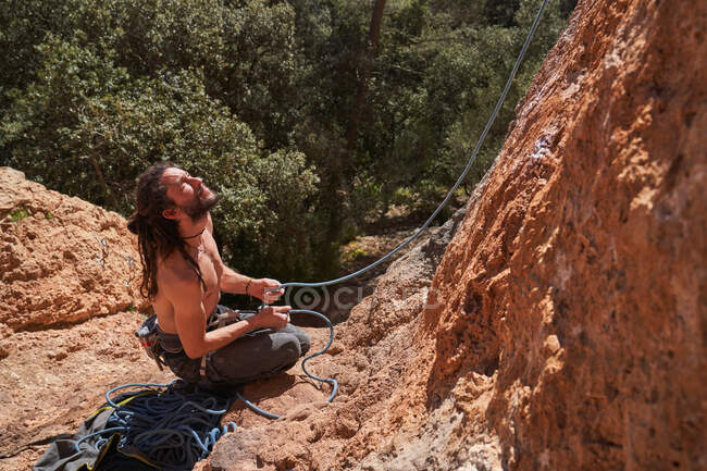 Сверху вид активного бородатого альпиниста, сидящего на краю скалы и держащего верёвку в руках, глядя вверх — стоковое фото