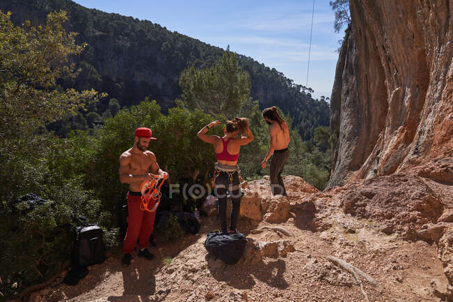 Group of professional climbers in colorful sportswear preparing equipment for ascending while standing next to rocky cliff in forest on sunny day — Stock Photo