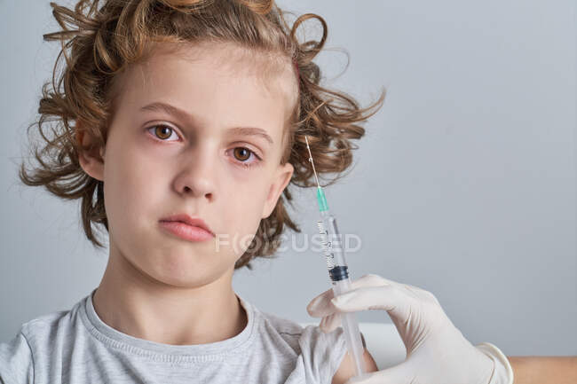 Неузнаваемый культуролог в латексных перчатках, заполняющий шприц вакциной из бутылки, готовящейся к инъекции в плечо мальчика с вьющимися волосами — стоковое фото