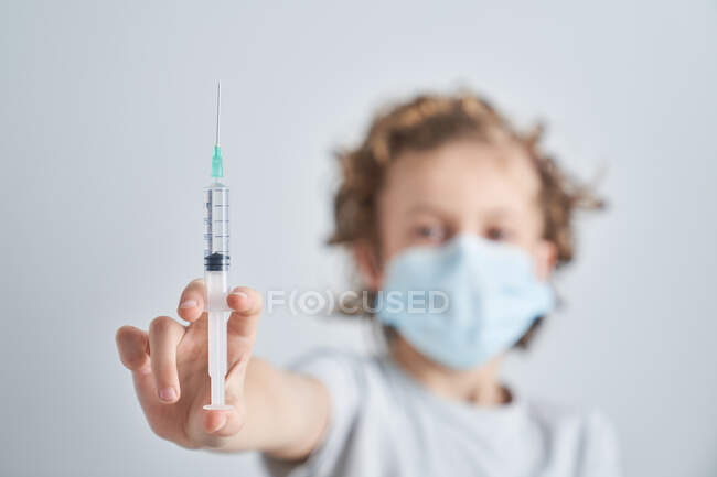 Menino irreconhecível com cabelo encaracolado e em máscara médica mostrando seringa com medicação vacinal enquanto visita o médico no hospital — Fotografia de Stock