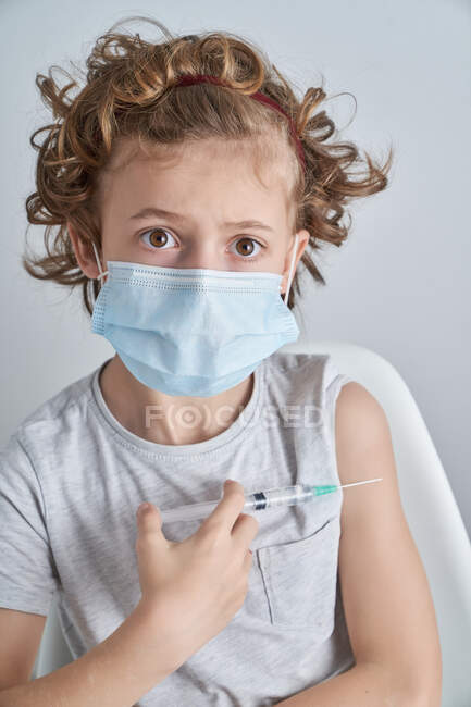 Niño rizado asustado con máscara médica sosteniendo la jeringa en la mano antes de administrarse la autoinyección de la vacuna en el hombro mirando a la cámara - foto de stock