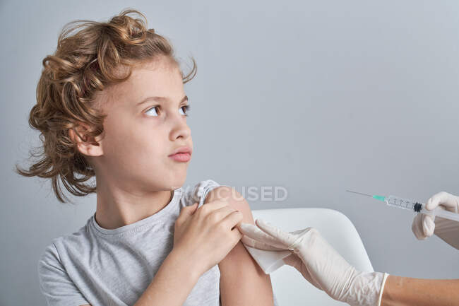 Crop nurse in white latex gloves holding shoulder of boy with curly hair while giving vaccine injection with syringe in modern clinic — Stock Photo