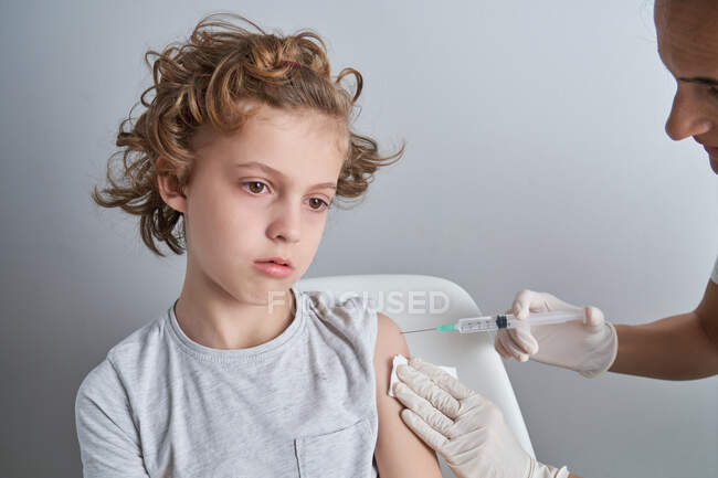 Crop nurse in white latex gloves holding shoulder of boy with curly hair while giving vaccine injection with syringe in modern clinic — Stock Photo
