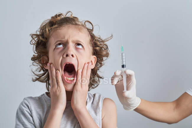 Кричущий маленький хлопчик з відкритим ротом торкається обличчя і дивиться вгору, а медсестра врожаю тримає шприц з ін'єкцією вакцини на сірому фоні — стокове фото