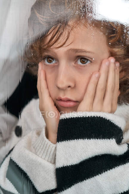 Menino entediado com as mãos nas bochechas olhando para a janela molhada enquanto descansa em casa no dia chuvoso — Fotografia de Stock
