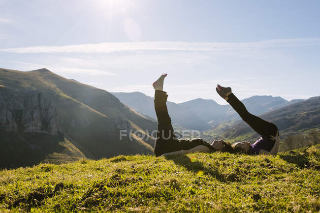 Vue latérale de la mère et de la fille en tenue de sport faisant de l'exercice avec les jambes levées tout en étant couchés ensemble sur l'herbe verte de prairie dans les montagnes ensoleillées en Cantabrie — Photo de stock