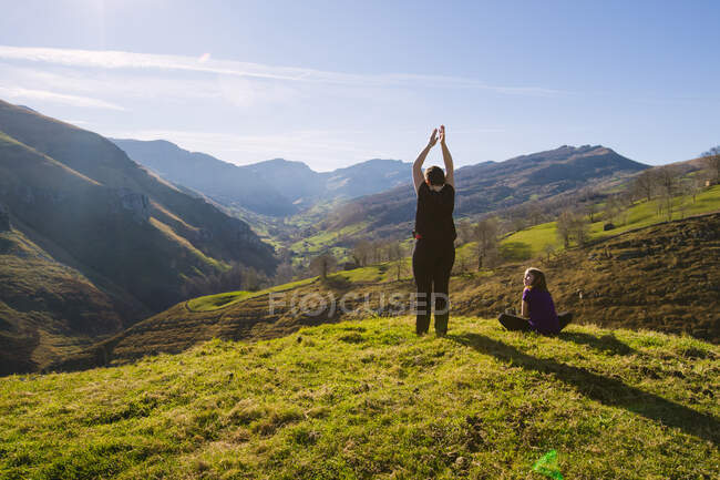 Погляд на анонімну жінку, яка стоїть з піднятими руками біля дочки, сидячи на зеленій траві під час прогулянки по сільській місцевості Іспанії. — стокове фото
