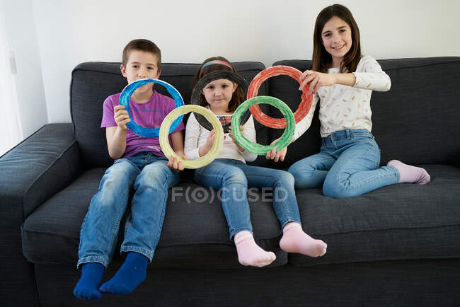 Group of playful children in casual jeans and shirts sitting on sofa and holding colorful disks in hands in shape of symbol of Olympic games — Stock Photo