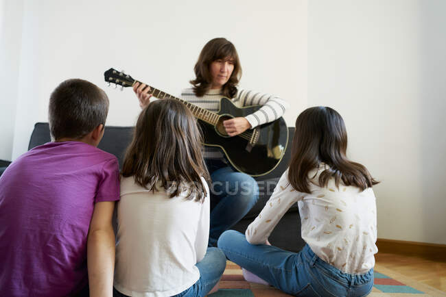 Mujer alegre tocando la guitarra para los niños en la sala de estar en casa - foto de stock