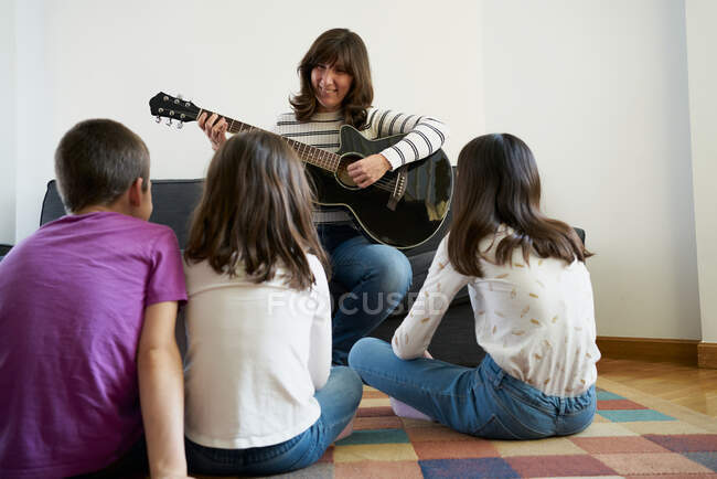 Mulher alegre em roupas casuais sentado no sofá confortável e tocando guitarra acústica para grupo de crianças sentadas no tapete no chão na acolhedora sala de estar — Fotografia de Stock