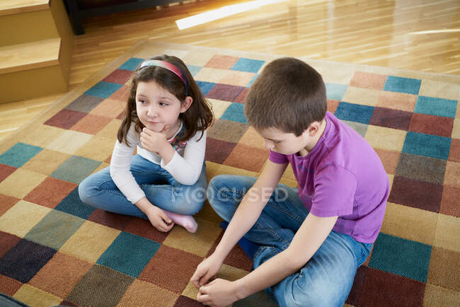 Desde arriba niño y niña en jeans casuales y camisas jugando juntos mientras están sentados con las piernas cruzadas en la alfombra en el suelo de madera - foto de stock