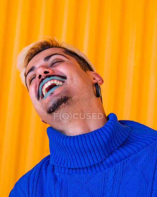 Андрогинный веселый мужчина с голубыми губами и в стильной одежде стоит на фоне желтой стены в городе с закрытыми глазами — стоковое фото