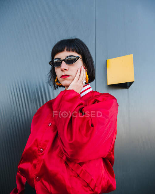 Feminino millennial vestindo jaqueta vermelha vívida e óculos de sol elegantes de pé com braços levantados na rua e olhando para a câmera — Fotografia de Stock