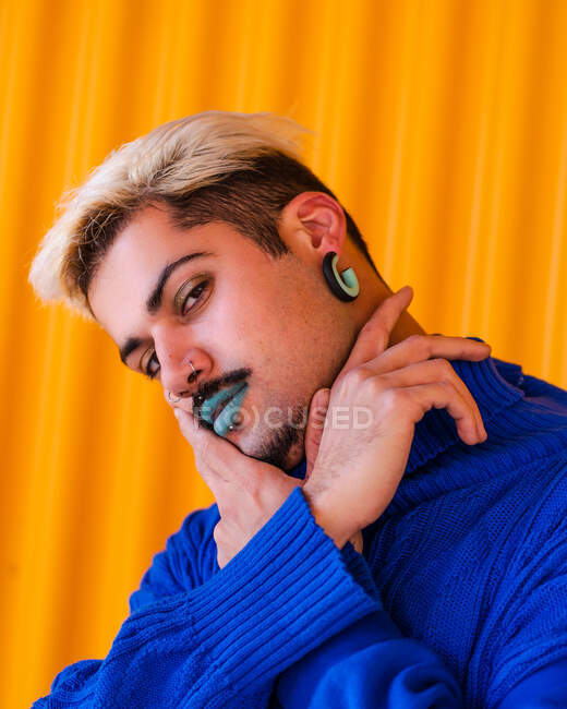Homem andrógeno com lábios azuis e em elegante desgaste em pé no fundo da parede amarela na cidade, enquanto olha para a câmera e rosto tocante — Fotografia de Stock