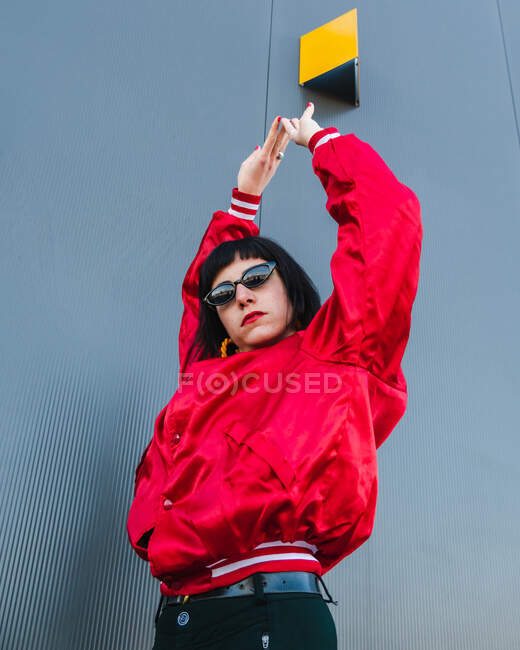 Bajo ángulo de mujer millennial vistiendo chaqueta roja vívida y gafas de sol con estilo de pie con los brazos levantados en la calle y mirando a la cámara - foto de stock