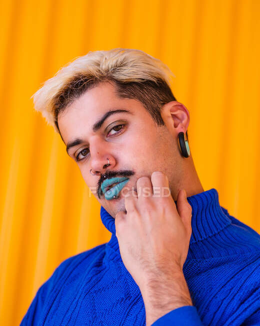 Maschio androgino con labbra blu e in elegante usura in piedi su sfondo di muro giallo in città, mentre guardando la fotocamera e toccando il viso — Foto stock