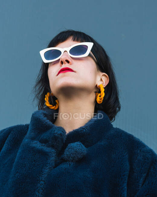 Von unten eine emotionslose Frau mit roten Lippen und in schickem Outfit, die in der Stadt vor einer Metallwand steht und mit stylischer Sonnenbrille nach oben schaut — Stockfoto