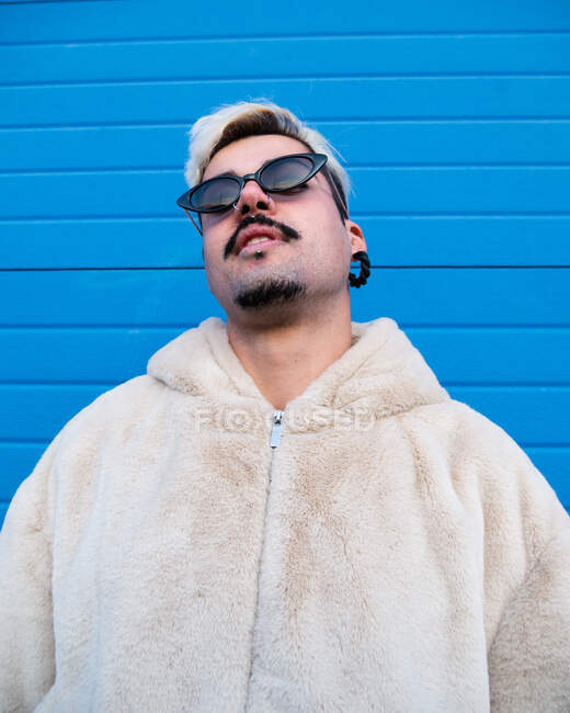 From below of determined cool male wearing stylish sunglasses standing in urban area on background of blue wall in city — Stock Photo