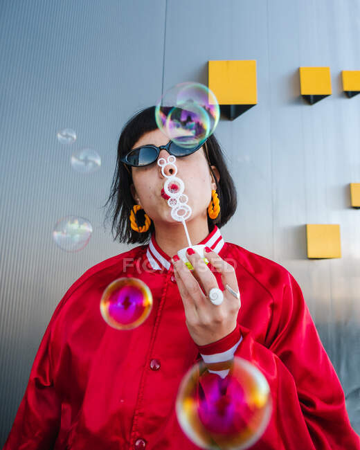 Young female in trendy cool clothes and sunglasses standing in city and blowing soap bubbles at camera — Stock Photo