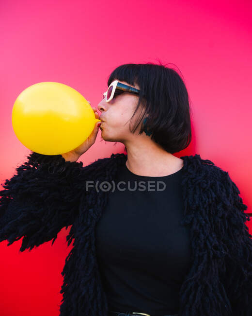 Elegante mujer soplando globo de aire amarillo sobre fondo rojo en la calle urbana y mirando hacia otro lado - foto de stock