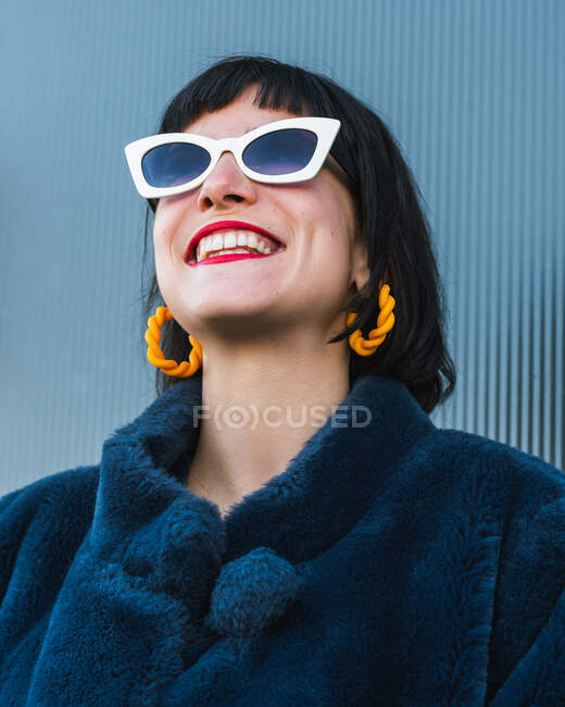 From below of delighted female with red lips and in fancy outfit standing against metal wall in city and looking up — Stock Photo