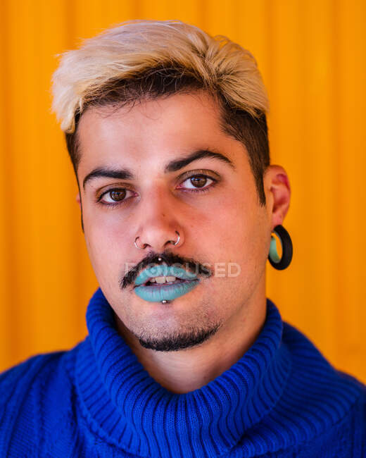 Homme androgyne aux lèvres bleues et à l'usure élégante debout sur le fond du mur jaune en ville tout en regardant la caméra — Photo de stock