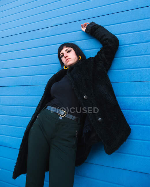 Unemotional female in fancy outfit standing against blue wooden wall in city and looking at camera — Stock Photo