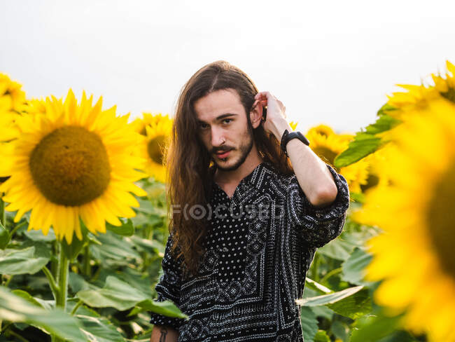 Sereno hipster masculino sin emociones con el pelo largo de pie en el campo de girasol amarillo y mirando a la cámara - foto de stock