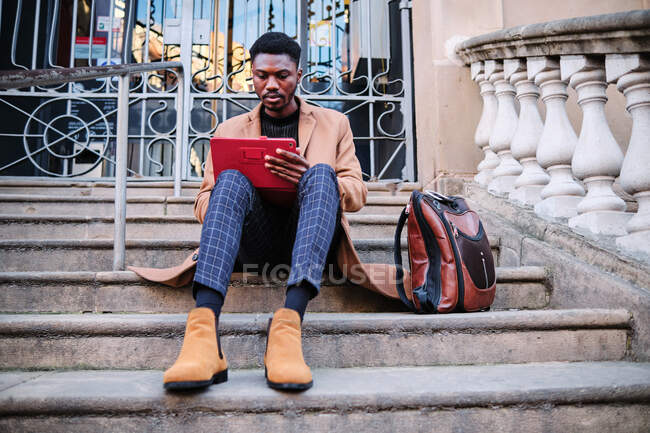 Современный чернокожий афроамериканец в стильной одежде и с серьгой на планшете, сидя на каменной лестнице на улице — стоковое фото