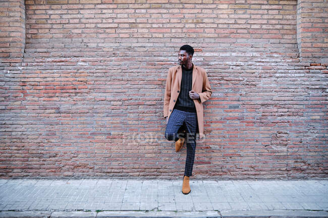 Moderno adulto negro afroamericano vestido con ropa elegante mirando hacia otro lado mientras está parado contra la pared de ladrillo de la calle - foto de stock