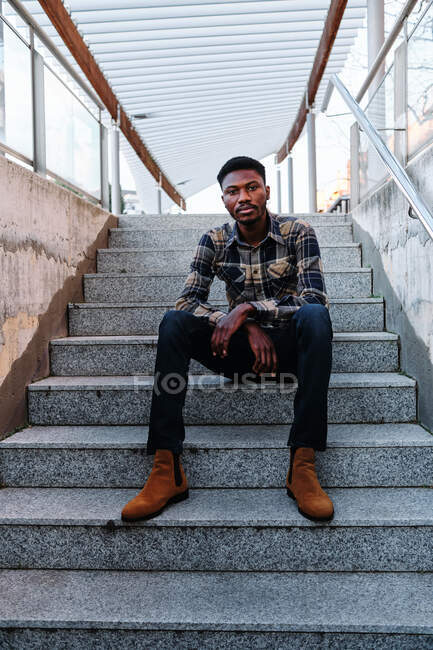 Moderno uomo afroamericano adulto non emotivo nero che indossa vestiti alla moda guardando la fotocamera mentre seduto nelle scale sulla strada della città — Foto stock
