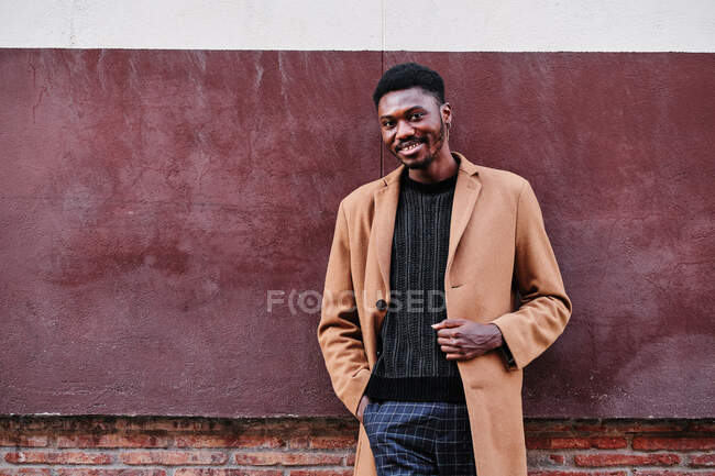 Moderno adulto contento negro afroamericano hombre con ropa elegante y pendiente mirando a la cámara mientras está de pie contra la pared colorida calle - foto de stock