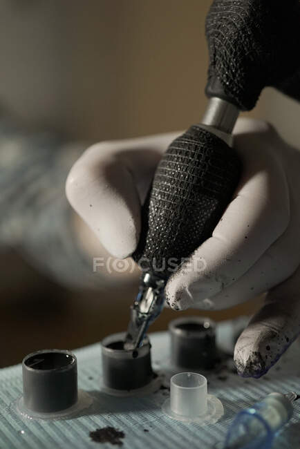 Крупним планом врожаю невизначений татуювач, який занурює татуювальну машину в чорне чорнило, працюючи в салоні — стокове фото