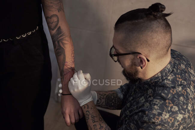 High angle of male tattooist making freehand tattoo and drawing sketch on arm of crop client in salon — Stock Photo