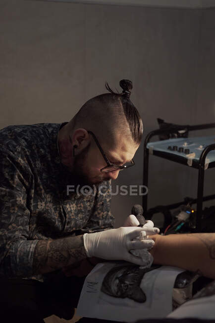 Tatuaje usando máquina y haciendo tatuaje con tinta negra en salón en cliente recortado anónimo - foto de stock