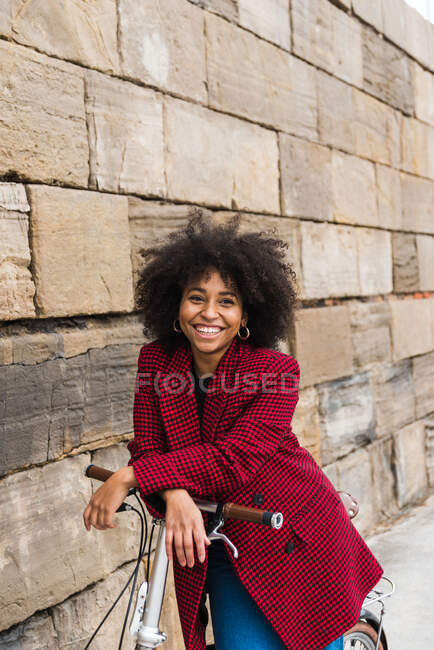 Вид спереду позитивної чорної жінки, що йде з велосипедом на вулиці вздовж старої кам'яної стіни і дивиться на камеру — стокове фото