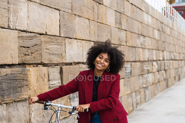 Vista frontal de hembra negra positiva caminando con bicicleta en la calle a lo largo de la vieja pared de piedra y mirando a la cámara - foto de stock