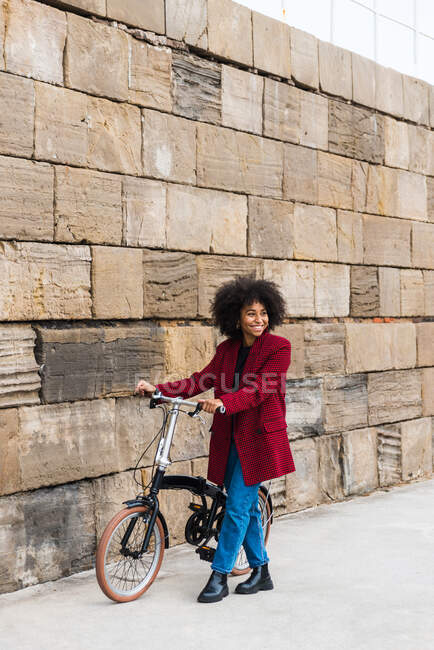 Front view of positive black female walking with bicycle on street along old stone wall and looking away — Stock Photo