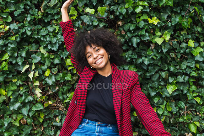 Happy black female with curly hair and raised arm enjoying weekend in park and looking at camera — Stock Photo