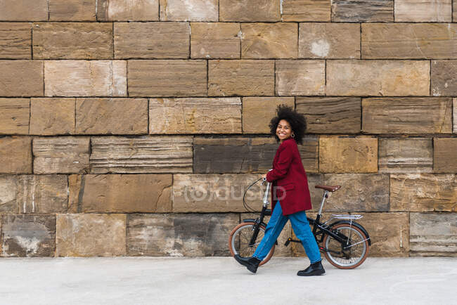 Vista laterale della positiva passeggiata femminile nera con bicicletta sulla strada lungo il vecchio muro di pietra e guardando la fotocamera — Foto stock
