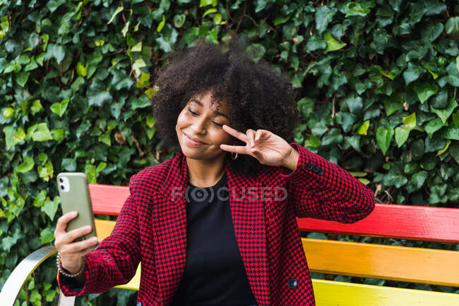 Content African American female sitting on bench in city and taking self portrait on smartphone while showing two fingers gesture — Stock Photo
