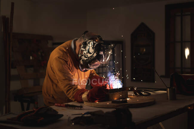 Unrecognizable male employee in protective gloves and helmet using welding machine while working in dark workshop — Stock Photo